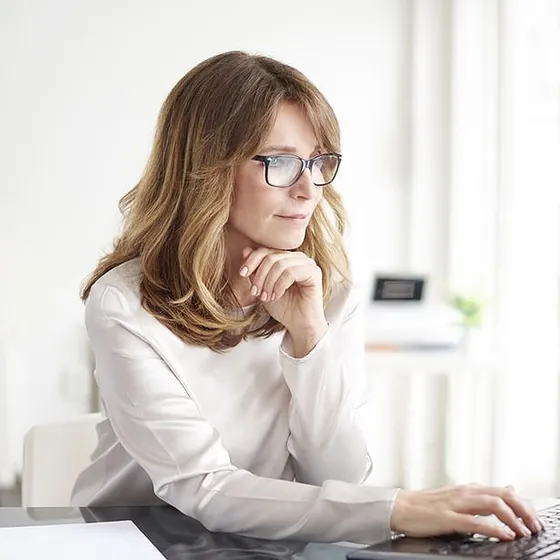 Woman in glasses resting her chin on her hand as she reads a laptop screen at a white desk