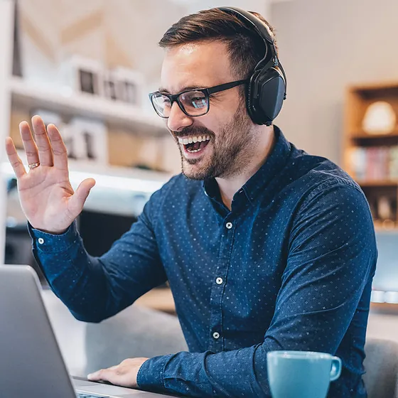 Smiling man in a headset waving at his laptop during a video call at a home office desk
