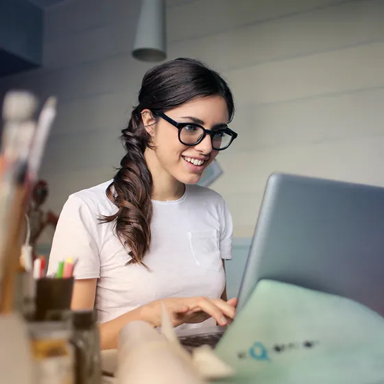 Woman in glasses smiling as she types on a laptop in a busy office