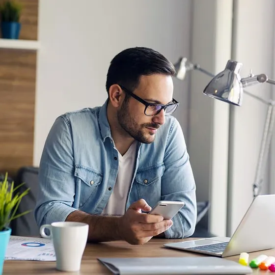 Man in glasses working on a laptop and phone at a sunny desk beside a desk lamp