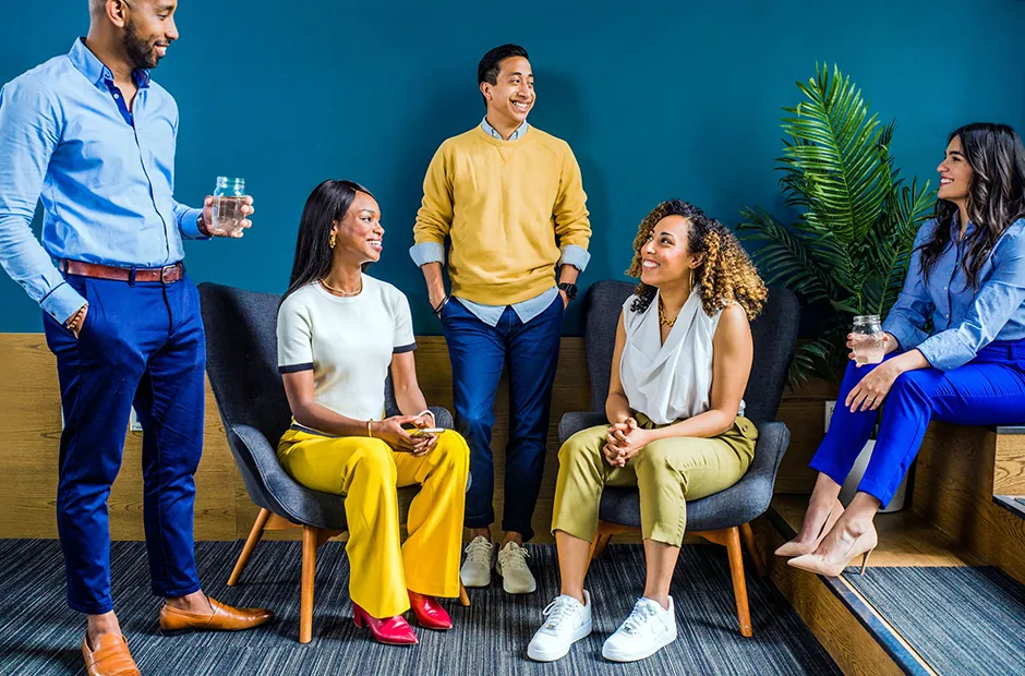 Five colleagues chatting and laughing in a lounge area with blue chairs against a teal wall