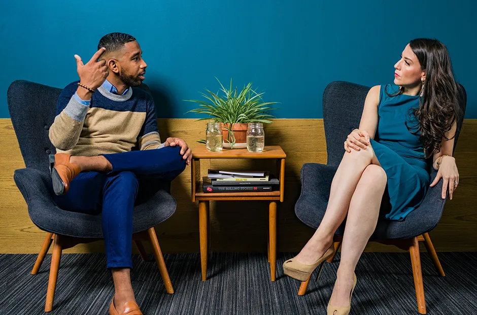 Two colleagues in conversation on blue armchairs in an office lounge with a teal wall and plants