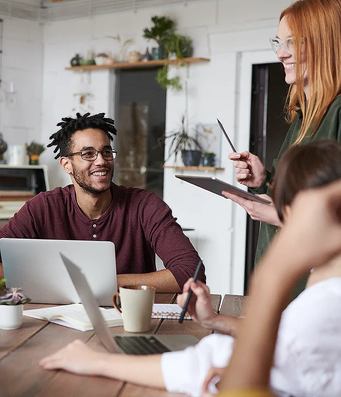 Team meeting around a laptop and coffee mugs, a smiling man in a maroon jumper and a colleague with a tablet
