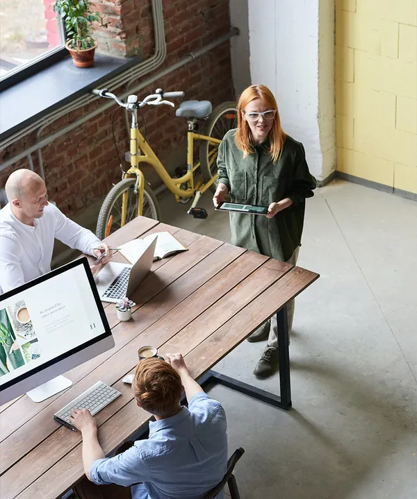 Colleagues working at laptops around a long wooden table in a bright open-plan office, a bicycle by the wall