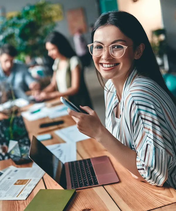 Smiling woman in glasses and a striped shirt holding a phone at her laptop, colleagues working behind her
