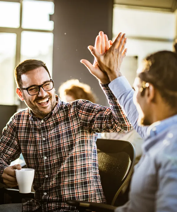 Two colleagues high-fiving across a desk in an office, one in a checked shirt holding a coffee mug