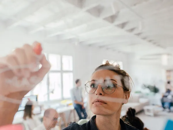 Woman in glasses writing on a glass board with a marker in a bright, airy office