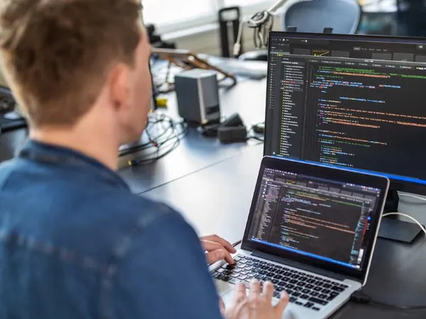 Developer in a denim shirt writing code on a laptop with a second screen of code beside him