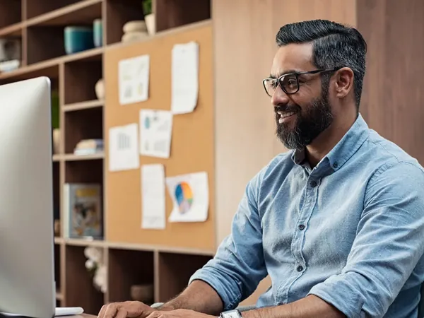 Bearded man in glasses smiling at his monitor at a desk, a pinboard of notes behind him